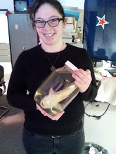 White girl with short curly black hair and glasses holding a fish in a jar